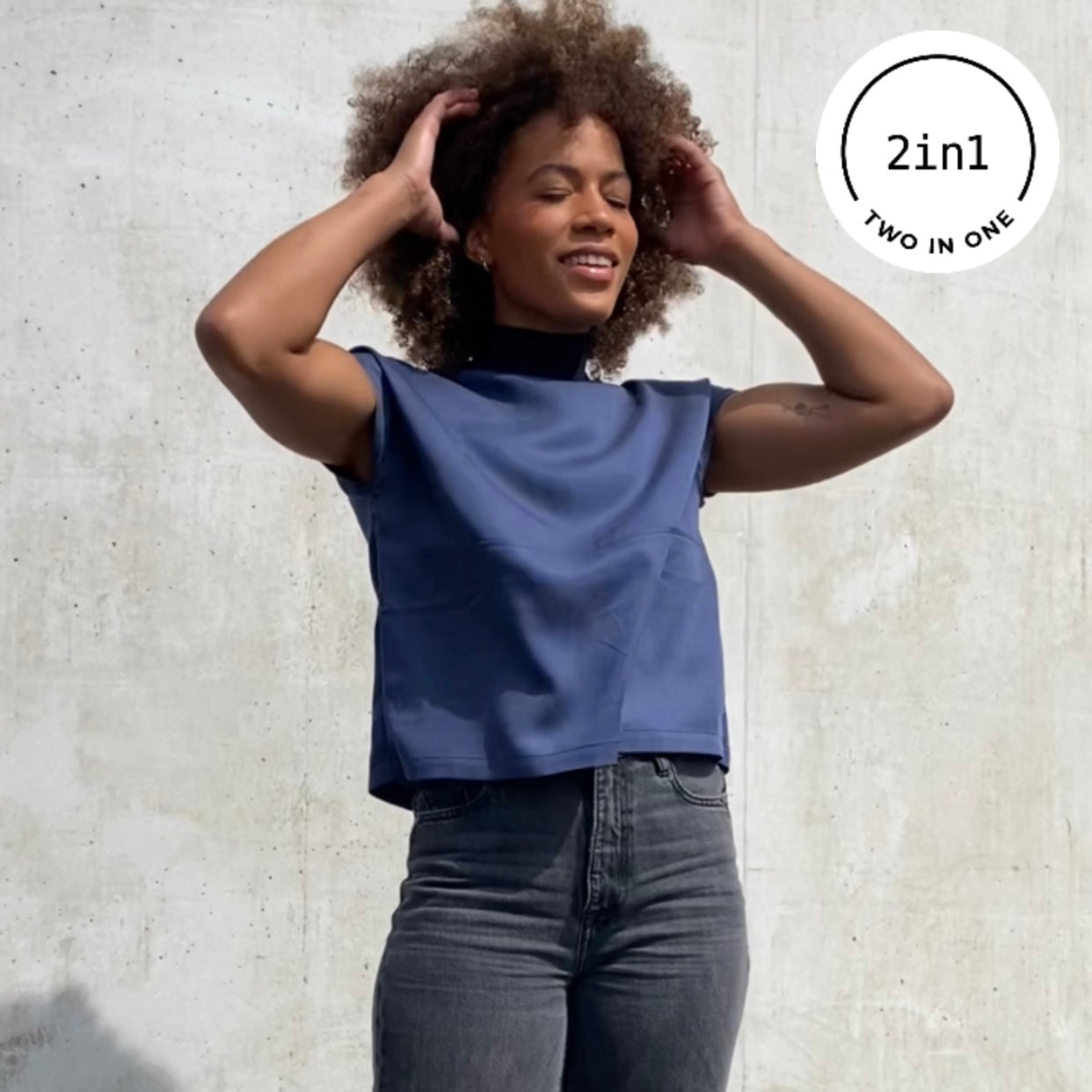 Frau mit indigo-farbener Bluse mit Reißverschluss vor grauer Betonwand mit 2IN1-Icon