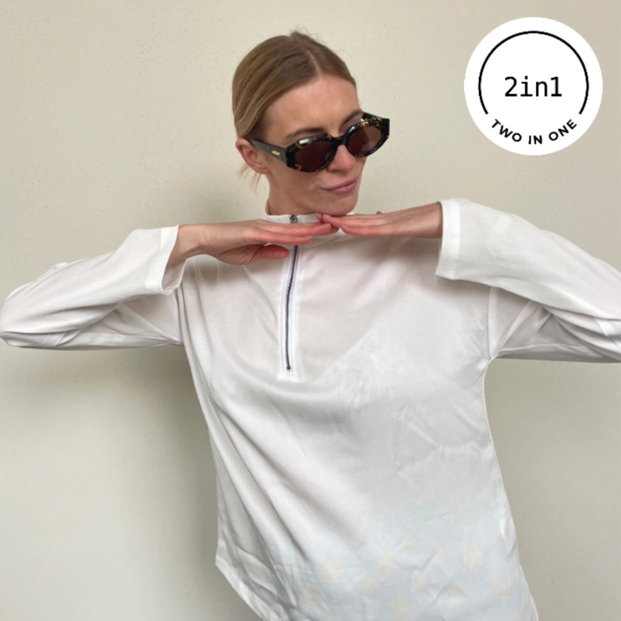 Blonde Frau mit weißer Bluse mit Reißverschluss und Sonnenbrille vor beigem Hintergrund mit weißem, rundem 2IN1-Icon