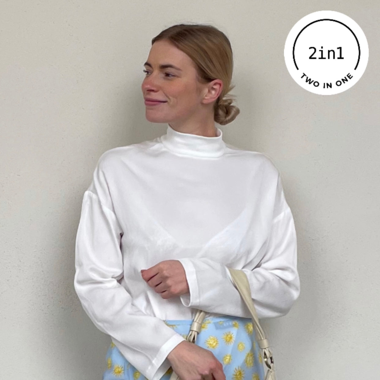 Blonde Frau mit weißer Bluse mit Stehkragen und Sonnenbrille vor beigem Hintergrund mit weißem, rundem 2IN1-Icon