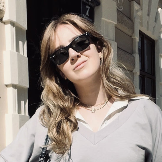 Gesicht einer Frau mit schwarzer Sonnenbrille und grauem Pulli vor beigen Häusern