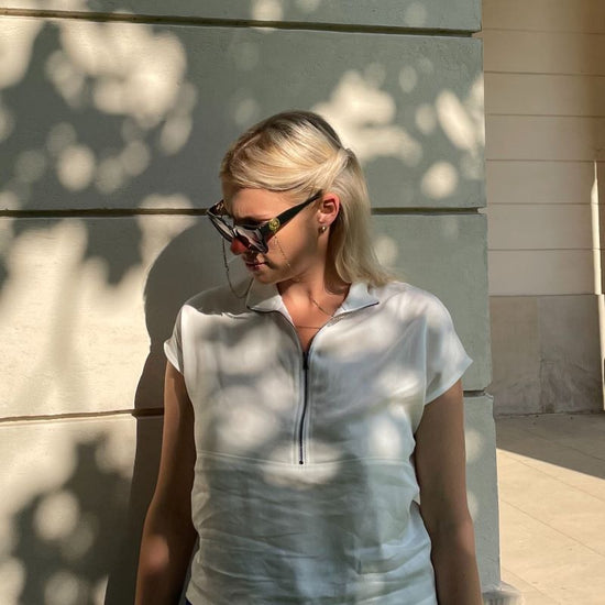 Oberköprper einer blonden Frau mit weißer Bluse vor beigem Hintergrund mit Schatten von Bäumen